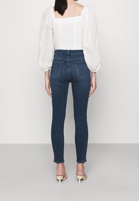Jeans skinny de talle alto, de color azul oscuro, hechos de denim, con un diseño ajustado y dos bolsillos traseros. Combinados con una blusa blanca texturizada.