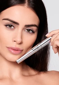 Produkt tuszu do rzęs w cylindrycznej, metalicznej srebrnej tubce z czerwono-czarnym logo. Zbliżenie na twarz kobiety trzymającej produkt.