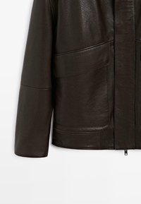 Giacca di pelle marrone scuro con una texture liscia, caratterizzata da un colletto rialzato, grandi tasche frontali e una chiusura con zip frontale.