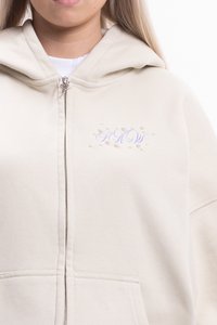 Béžová mikina na zips s kapucňou, na ktorej je nápis "HOW" v levandulovej farbe, ozdobená dekoratívnymi korálkami, z mäkkého materiálu, vo voľnom strihu.