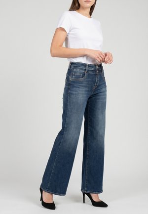 Mujer que lleva una camiseta blanca, vaqueros azules de pierna ancha y talle alto, y zapatos de tacón negros, de pie frente a un fondo liso.