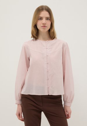 Mujer de frente, con blusa rosa claro de manga larga con botones y pantalón marrón, de pie contra un fondo liso.
