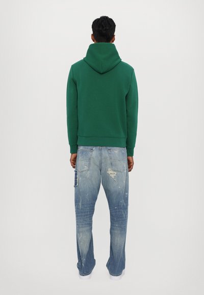 Felpa verde con cappuccio ampio, polsini a coste e vestibilità rilassata. Abbinata a jeans blu chiaro con effetti usurati e tasca laterale.