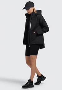 Giacca nera impermeabile con cappuccio, cerniera e tasche frontali; abbinata a pantaloni corti neri e sneakers; design minimalista e texture liscia.