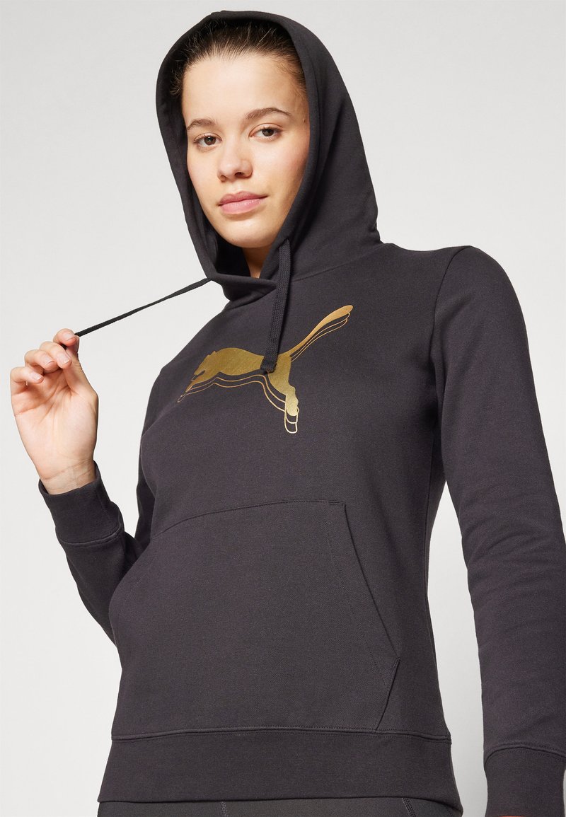 Schwarzer Kapuzenpullover aus weichem Material mit einem goldenen Puma-Logo auf der Vorderseite und einer frontalen Tasche. Eng anliegende Passform mit gerippten Bündchen.