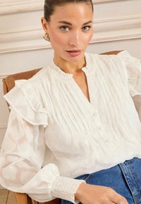 Bílá plisovaná blůza s řasenými rukávy, s knoflíkovým zapínáním a texturovanou tkaninou, kombinovaná s modrými džíny. Elegantní a vzdušný design.