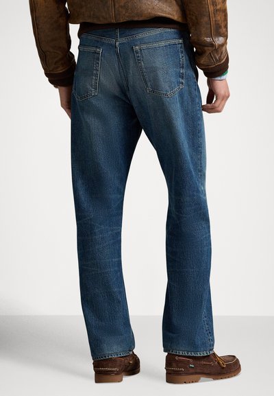 Σκούρο μπλε τζιν παντελόνι με ίσια γραμμή, με πίσω τσέπες και διακριτική ξεθώριασμα, συνδυασμένο με καφέ παπούτσια στυλ μοκασίνι.