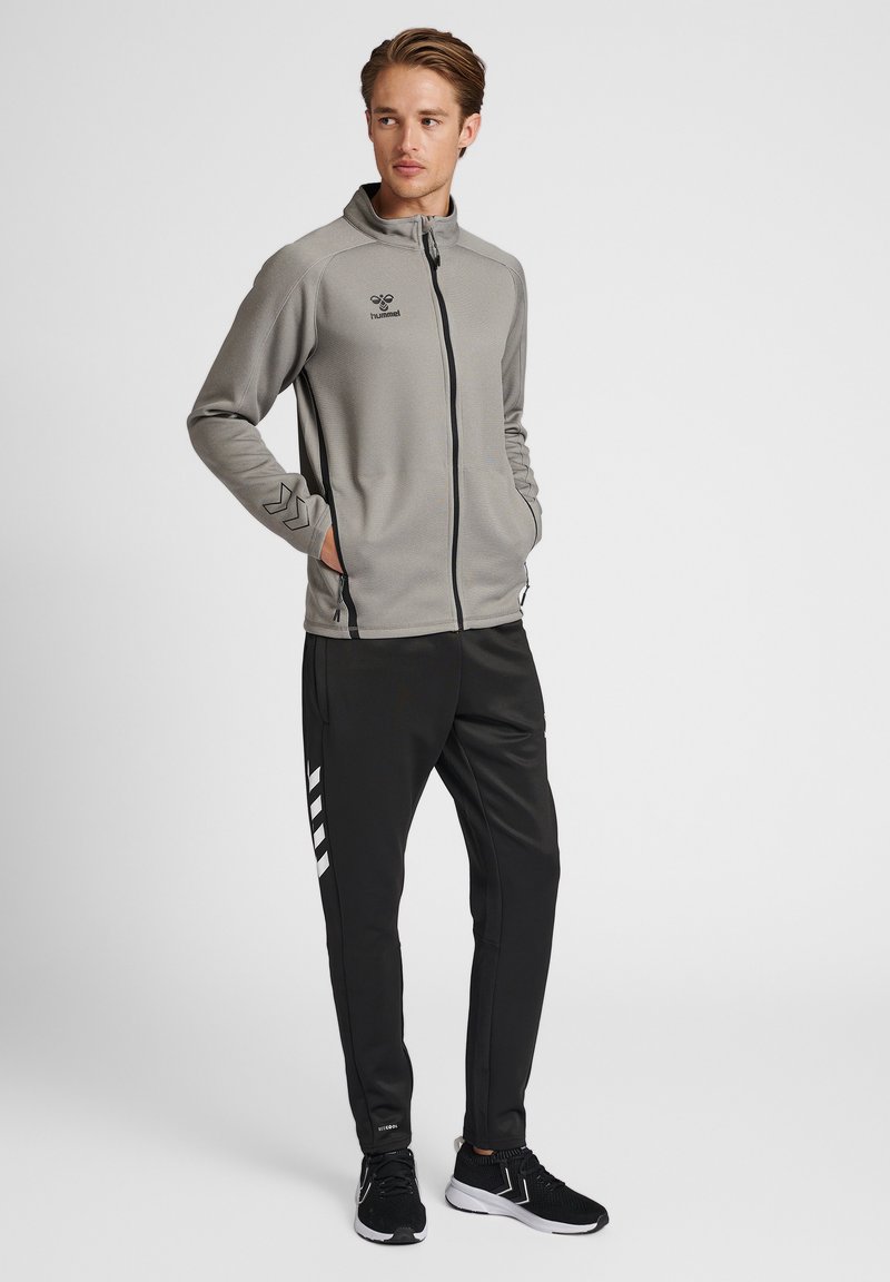 Graue Zip-Jacke mit schwarzem Logo, kombiniert mit schwarzen Sporthosen mit weißen diagonalen Streifen, getragen mit schwarzen Sneakers.