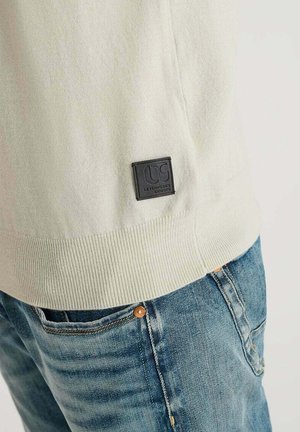 Pull crème clair avec un ourlet côtelé, comportant un patch logo noir sur le côté. Associé à un jean en denim bleu délavé.