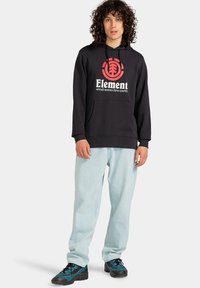 Sort hættebluse med hvid "Element" logo og rød grafik, parret med lyseblå jeans og sorte og blå sneakers.