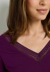 Haut violet à col en V mettant en valeur un délicat motif en dentelle le long de l'encolure. Le tissu semble lisse avec une coupe légèrement décontractée.