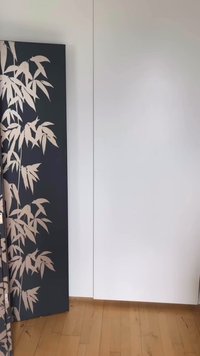 Svart sammenleggbart skjerm med beige bambusbladmønster som lener mot en hvit vegg på et tregulv.