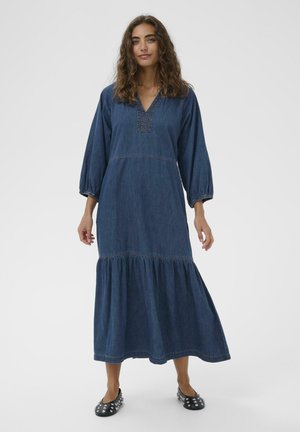 Kobieta z kręconymi włosami, ubrana w długą niebieską sukienkę z denimu z marszczonym dołem i balonowymi rękawami, stojąca na jednolitym jasnym tle.