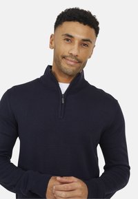 Felpa con zip in navy in un morbido tessuto a maglia, con colletto alto, maniche lunghe e una vestibilità aderente. I dettagli notevoli includono una cerniera in metallo.