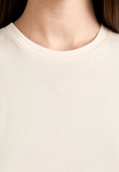 Close-up of a person's neck and upper chest wearing a plain beige round-neck shirt, with long brown hair visible.