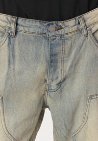 Jeanși din denim de culoare albastru deschis cu o textură decolorată, având închidere cu buton și cusături portocalii contrastante. Buclele sunt vizibile pe laturi.