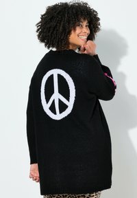 Sort strikket sweater med stort hvidt fredssymbol på ryggen. Har lyserøde detaljer på ærmerne og en løs pasform.