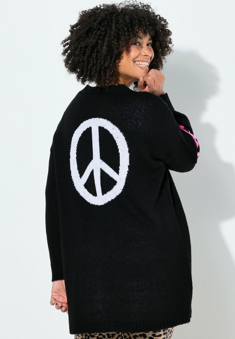 Sort strikket sweater med stort hvidt fredssymbol på ryggen. Har lyserøde detaljer på ærmerne og en løs pasform.