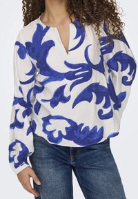 Blusa bianca con ampi motivi floreali blu, con scollatura a V e maniche lunghe a sbuffo, abbinata a jeans in denim.