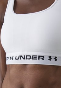 Närmre bild av en person som bär en vit sport-bh med "UNDER ARMOUR" och logotypen på det elastiska bandet under bysten.