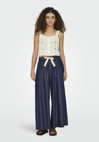 Bílý háčkovaný crop top s knoflíkovým zapínáním, doplněný širokými tmavými džíny se světle béžovým zavazovacím páskem. Celý vzhled doplňují ploché sandály.
