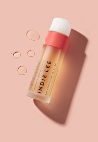 Bottle of Indie Lee AHA exfoliating solution with coral cap, placed on peach background with droplets of liquid nearby.