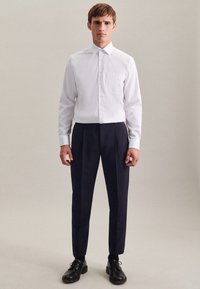 Weißes Button-Up-Hemd mit einer Brusttasche, kombiniert mit marineblauen, gepleated Hosen und schwarzen Lederschuhen, auf einem Modell vor einem neutralen Hintergrund.