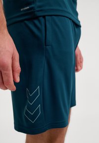 Pantalones cortos atléticos en color verde azulado, hechos de un material transpirable, con un patrón texturizado y un acento reflectante en forma de chevrón en el lado inferior.