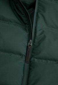 Ciemnozielona kurtka puchowa z pełnym zamkiem błyskawicznym. Posiada pikowaną teksturę, etykietę z napisem na zamku oraz gładki materiał zewnętrzny.