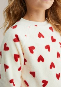 Hanorac alb pufos cu textură moale, având modele în formă de inimă roșie și design cu guler rotund, guler și mâneci raglan cu tivuri striate.