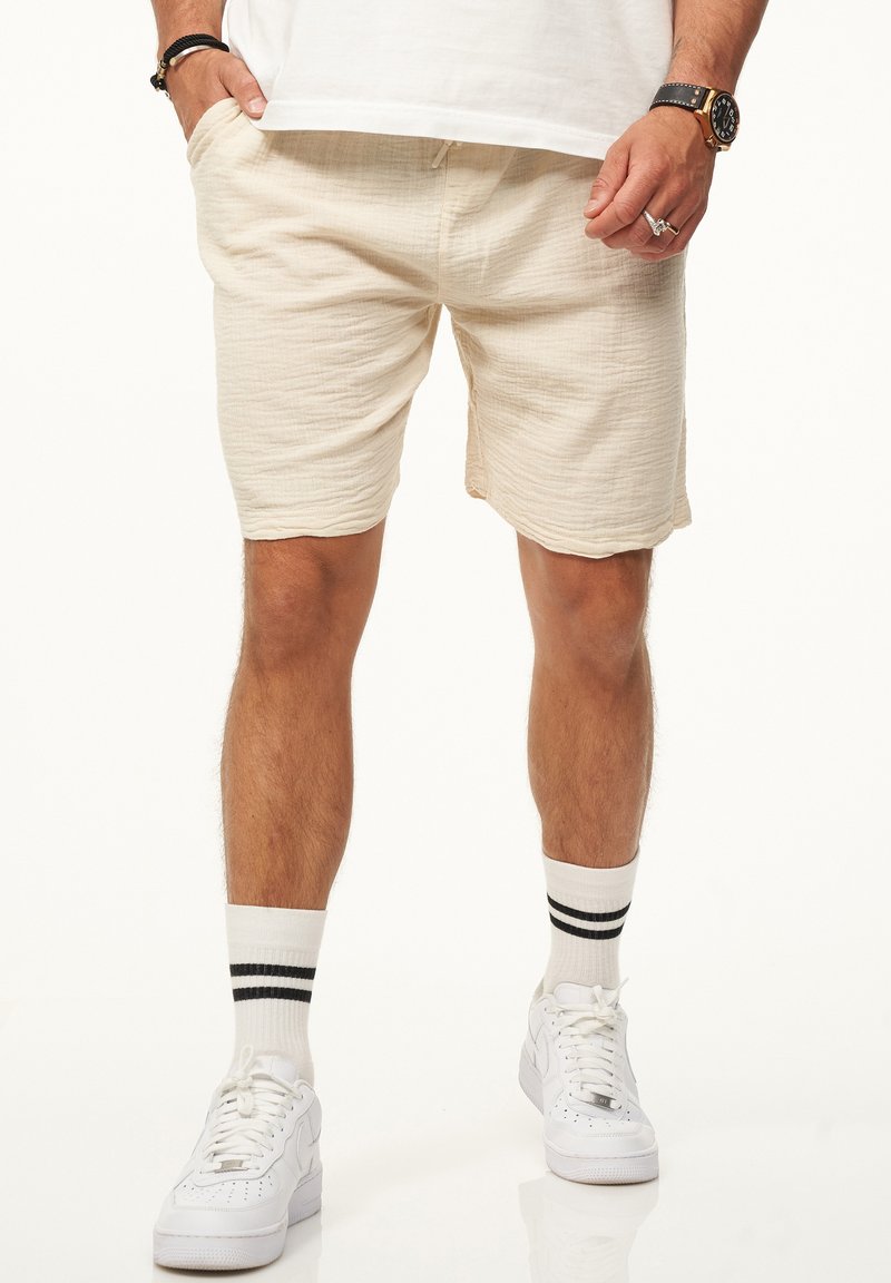 Beige Textur-Shorts mit elastischem Bund und seitlichen Taschen. Getragen mit weißen Sneakers und gestreiften weißen Socken.