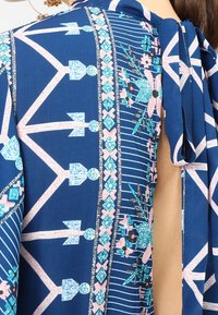 Modrý materiál s geometrickými tvary, květinovými vzory a uvázanou mašlí na rameni ženy, která odhaluje část pokožky na zádech.