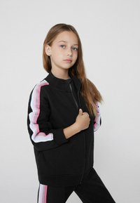 Giacca nera con zip e maniche lunghe, caratterizzata da strisce rosa e azzurro chiaro lungo le maniche. Tessuto morbido con colletto e polsini a costine.