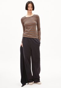 Langärmliges, braun- und marineblau gestreiftes Oberteil, kombiniert mit dunkelblauen, locker sitzenden Jogginghosen. Das Outfit beinhaltet einen dunklen Pullover, der über einem Arm hängt.