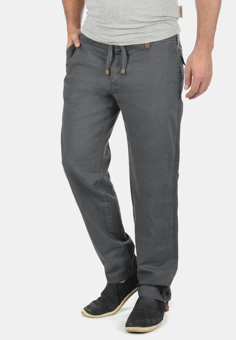 Pantalon gris en tissu léger avec une coupe décontractée, taille à cordon, poches latérales et revers roulés, associé à des chaussures noires.