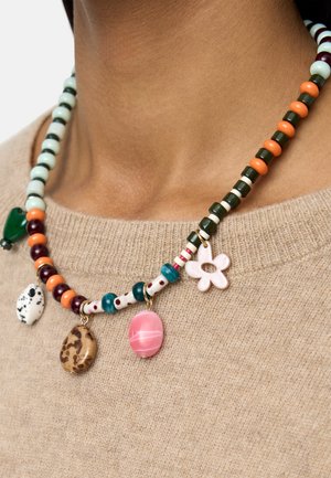 Naszyjnik z wielokolorowymi koralikami i pięcioma zawieszkami, w tym różowym owalem, białym kwiatem oraz wzorami przypominającymi kamienie, na beżowym swetrze.