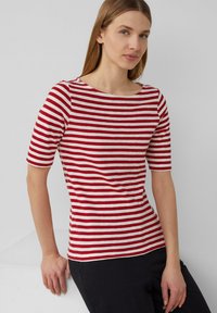 T-shirt rayé rouge et blanc avec un col bateau et des manches courtes. Tissu doux avec un design ajusté, associé à un pantalon foncé.