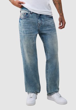 Mann trägt ausgebleichte blaue Jeans mit geradem Bein, weiße Sneakers und ein weißes Hemd mit sichtbaren tätowierten Armen.
