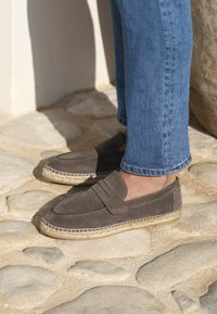 Mocassins en daim gris avec une semelle en jute tressé, comportant un accent cousu sur le dessus, associés à un jean en denim bleu sur une surface en pierre.