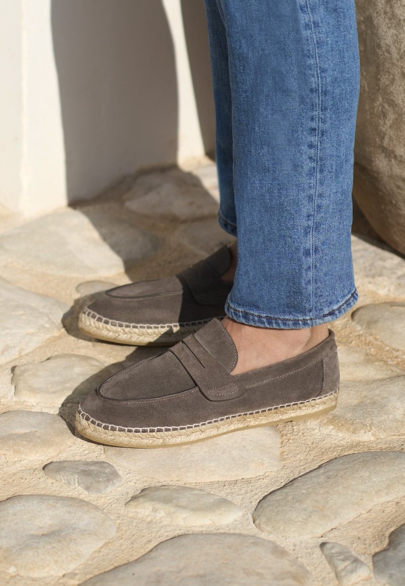 Mocassins en daim gris avec une semelle en jute tressé, comportant un accent cousu sur le dessus, associés à un jean en denim bleu sur une surface en pierre.