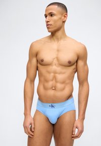 Μυώδης άνδρας που φοράει ανοιχτό μπλε ανδρικά εσώρουχα Calvin Klein, στέκεται με τα χέρια χαλαρά, κοιτάζοντας στο πλάι μπροστά σε απλό λευκό φόντο.