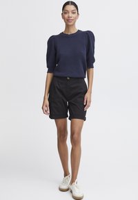 Kvinna i en strukturerad marinblå kortärmad topp, svarta uppvikta shorts och vita sneakers, stående mot en enkel vit bakgrund.