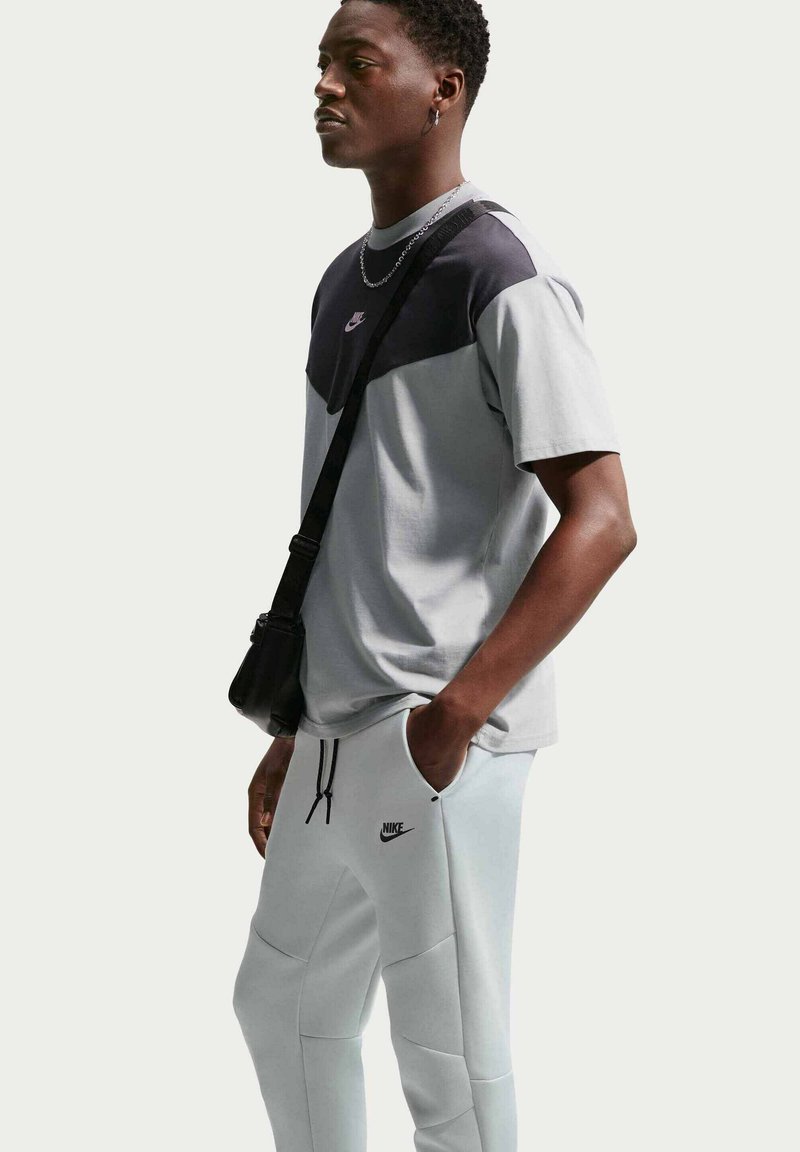 Junger Mann trägt ein graues und schwarzes Nike-T-Shirt und hellgraue Nike-Sweatpants, trägt eine schwarze Umhängetasche und steht seitlich.