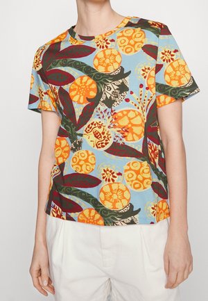 Personne portant une chemise à manches courtes avec un motif floral abstrait coloré en orange, rouge, vert et bleu, associée à un pantalon blanc.