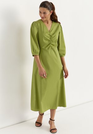 Vrouw in een groene midi-jurk met geplooide voorkant en opgeblazen driekwartmouwen, draagt zwarte sandalen met enkelbandje en ringen, staand tegen een witte muur.