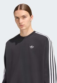 Μαύρο φούτερ με λευκή τριπλή ρίγα στα μανίκια και μικρό γκρι λογότυπο στο στήθος. Απαλό ύφασμα, άνετη εφαρμογή.