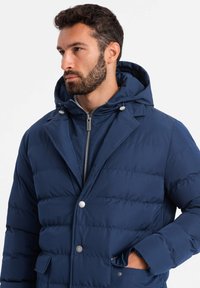 Chaqueta de plumas azul marino con capucha, con costuras horizontales, botones a presión plateados y cremallera en la parte delantera, fabricada en un tejido suave y duradero.