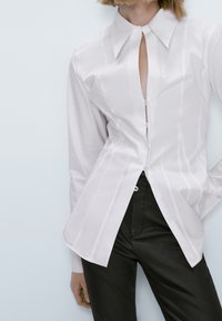 Vit skjorta med knappar och en uringning vid bröstet, långa ärmar och vertikala ränder. Kombination med svarta, figursydda byxor.