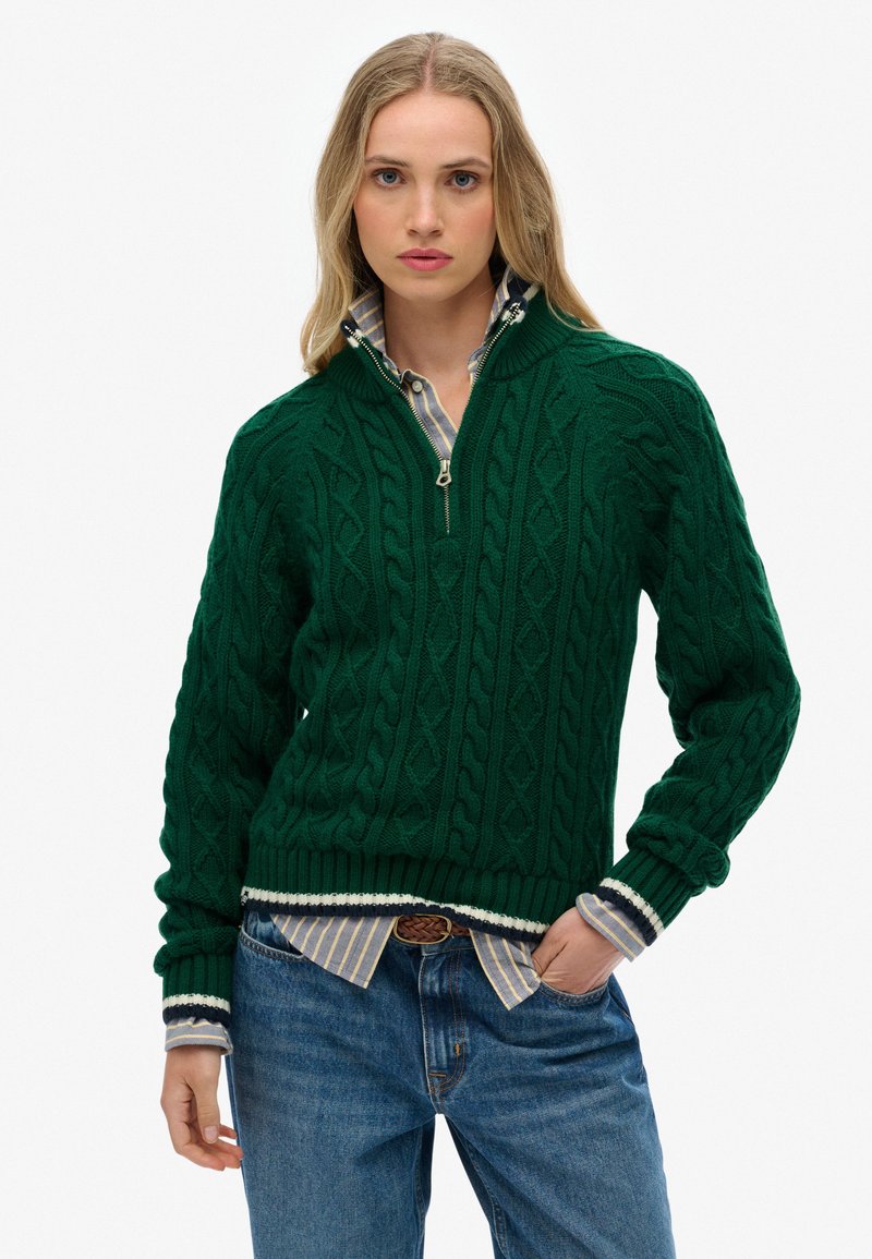 Grüner Strickpullover mit einem Zopfmuster, kurzem Reißverschluss und gerippten Bündchen. Getragen über einem karierten Hemd mit Kragen und blauen Jeans.
