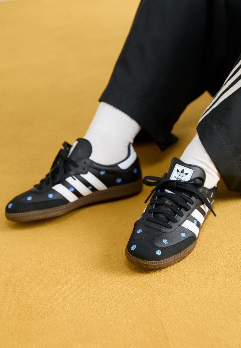 Black sneakers with white stripes, blue floral accents, rubber sole, and black laces. Worn with white socks and black pants on a yellow background.